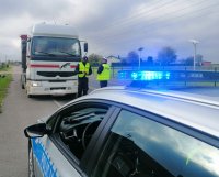na pierwszym planie oznakowany radiowóz piotrkowskiej drogówki, w tle policjanci kontrolujący samochód ciężarowy podczas akcji,,TIR&quot;