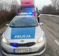 na pierwszym planie widoczny oznakowany radiowóz, w tle stojący policjant przy kabinie pojazdu ciężarowego podczas kontroli