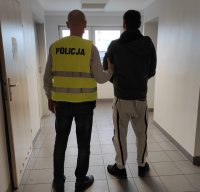 policjant po cywilnemu, w kamizelce odblaskowej z napisem policja prowadzi zatrzymanego przez korytarz