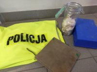 na zdjęciu widać policyjną kamizelkę,a przy niej słoik z dilerkami , notatnik, plastikową niebieską apteczkę zabezpieczone przez policjantów