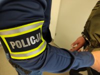 Opaska z napisem policja na rękawie policjanta po prawej ręce zatrzymanego w kajdankach