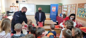 policjant oraz przedstawiciele Starostwa Powiatowego przekazują uczniom klas pierwszych odblaskowe kamizelki oraz książeczki z zasadami bezpieczeństwa