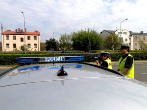 na zdjęciu dwoje policjantów ruchu drogowego mierzą prędkość , na pierwszym planie policyjny radiowóz