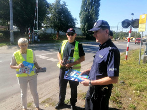 policjant trzyma zeszyty edukacyjne dedykowane dzieciom w wieku szkolnym, z  informacjami o bezpiecznych zachowaniach  w rejonie przejazdów kolejowych. Na zdjęciu wspólnie z przedstawicielami SOK