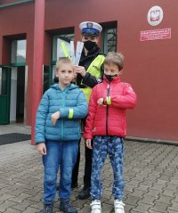 policjantka z uczniami przed budynkiem szkoły podstawowej podczas policyjnej akcji rozdawania odblasków