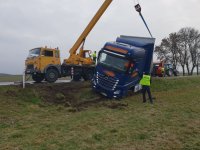 policjant stojący przy kabinie pojazdu ciężarowego, który zjechał z jezdni do rowu melioracyjnego, w tle widać dźwig wyciągający ciężarówkę