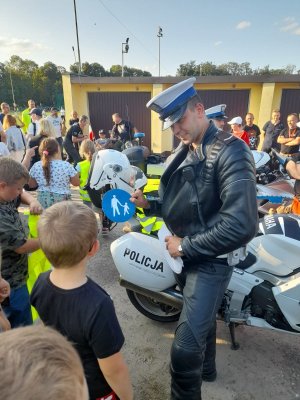 policjant ruchu drogowego podczas festynu miejskiego stoi obok policyjnego motocykla i pokazuje dzieciom znaki drogowe