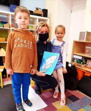 na zdjęciu dzielnicowa kuca  trzymając na kolanach dziewczynkę z grupy przedszkolnej, obok nich chłopiec