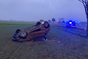 pojazd peugeot 206 przewrócony na polu w tle radiowóz policyjny z włączonymi sygnałami świetlnymi