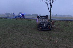pojazd peugeot 206 przewrócony na polu w tle radiowóz policyjny z włączonymi sygnałami świetlnymi