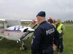 na zdjęciu widać policjanta stojącego przodem do uszkodzonej awionetki. Na drugim planie funkcjonariusze straży pożarnej