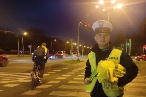 umundurowany policjant stojący przy przejściu dla pieszych, trzymający elementy odblaskowe, w tle widoczny rowerzysta z kamizelką odblaskową