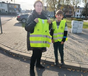 kobieta wraz z dzieckiem, kobieta przed sobą trzyma kamizelkę odblaskową, chłopiec stoi po prawej stronie i ma już założoną kamizelkę