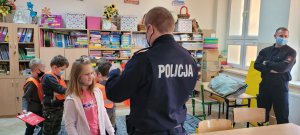 na zdjęciu policjant pokazuje uczennicy pierwszej klasy podstawówki jak prawidłowo nosic elementy odblaskowe