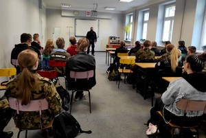 uczniowie siedzący w ławkach podczas spotkania z policjantem, w tle widoczny policjant prowadzący pogadankę