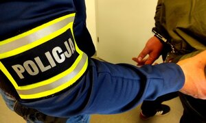 policyjna opaska, obok ręce zatrzymanego w kajdankach