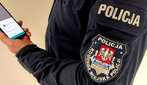 Policjant stojący z telefonem w ręku. Na wyświetlaczu widoczny jest kod blik.