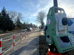 Urządzenie do wykonywania pomiarów podczas obsługi wypadków drogowych, w tle widoczny radiowóz i pojazd biorący udział w zdarzeniu.