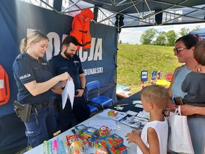 Policjant i policjantka pod namiotem na stole różne ulotki, malowanki, odblaski, przed stołem dzieci , mama z dzieckiem na ręku.