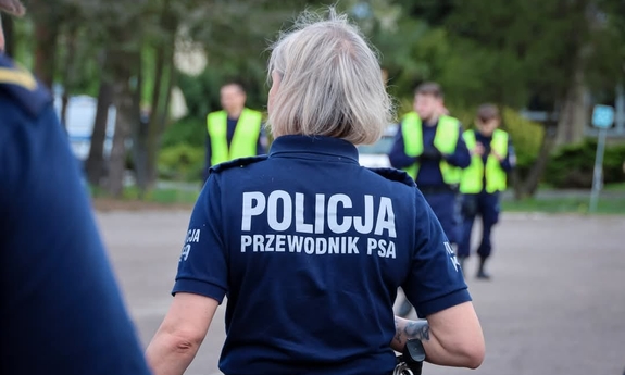 Policjantka w koszulce z napisem Policjant Prewodnik psa, stojąca tylem, w tle policjanci nieumundurowani w kamizelkach odblaskowych.